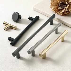 May include: A collection of cabinet hardware in various finishes. Includes a black knob, a gold T-shaped pull, and several bar pulls in black, gray, and gold. The hardware is arranged on a white surface, with a dried flower in the background.