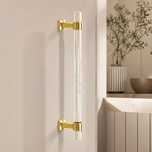 May include: A vertical door handle featuring a clear, cylindrical acrylic bar held by two polished gold-tone metal brackets. The handle is mounted on a flat surface, showcasing a sleek, modern design with metallic accents and transparent material.