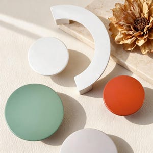 May include: A selection of contemporary cabinet hardware. It features a white, semi-circular handle and round knobs in white, green, and orange. The hardware is displayed on a light surface, suitable for home decor.