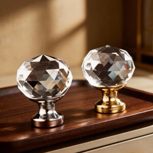 May include: Two crystal cabinet knobs with faceted surfaces. One knob has a silver base, and the other has a gold base. The knobs are displayed on a dark brown wooden tray, showcasing their decorative design.