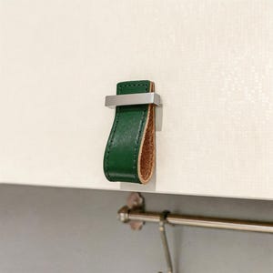 May include: A green leather cabinet pull with a stainless steel bar. The pull is attached to a white cabinet door. The interior of the pull is a light brown color. The cabinet pull is a modern design.
