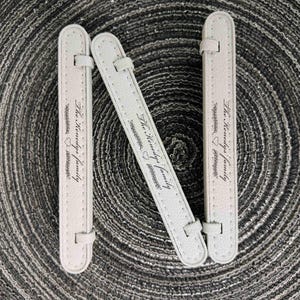 Puede incluir: Tres correas rectangulares de piel sintética texturizada en color blanco roto con extremos redondeados. Cada correa tiene una pequeña trabilla en la parte superior e inferior, y está grabada con el texto "The Kenedy's family" junto a un corazón y un gráfico floral.