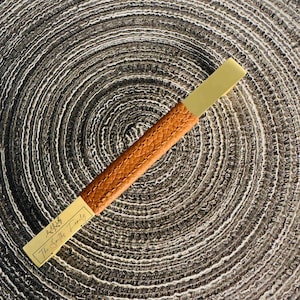 Puede incluir: Un tirador de cajón rectangular con una empuñadura central de cuero color canela texturizado y dos extremos de metal dorado. Un extremo está grabado con un pequeño icono de hoja y el texto The Smiths Family. El herraje tiene costuras visibles.