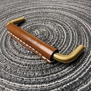 May include: A brass-toned metal cabinet handle wrapped in a smooth brown leather sleeve. The leather is secured with white stitching along the underside and features a rectangular logo stamp with the text The Smith Family and a leaf graphic.