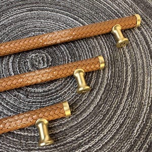 May include: Three cabinet pulls featuring a tan braided leather texture with gold-toned metal end caps and mounting posts. Each handle displays the white printed text The Luebbes Home with decorative flourishes on the leather surface.