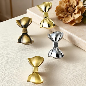 Pode incluir: Quatro puxadores decorativos para armários com acabamentos dourado, bronze e prateado. Cada puxador tem formato de laço com haste central e extremidades alargadas. Os puxadores são exibidos em uma superfície branca com fundo neutro.