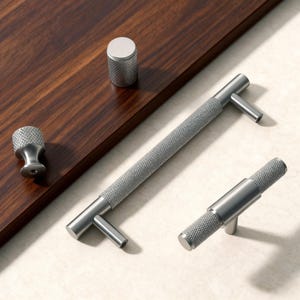 May include: A collection of silver cabinet hardware, including a knob, a cylindrical pull, and two T-bar pulls. The hardware has a textured, diamond-patterned surface and a brushed finish. The items are displayed on a dark wood surface.