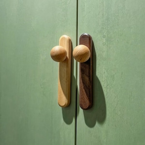 Op de afbeelding: Twee houten kastgrepen. Eén is licht van kleur, de andere is donkerbruin. Elke greep heeft een afgeronde knop aan de bovenkant en een rechthoekige basis. De grepen zijn gemonteerd op een groene kastdeur.
