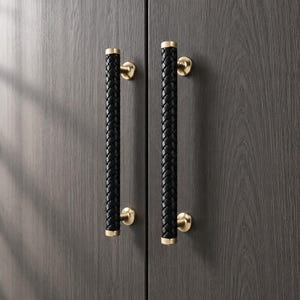 May include: Two vertical cabinet handles featuring a black braided texture wrapped around a cylindrical core. Each handle is secured to the dark wood cabinet doors by two circular, brushed gold metal mounting brackets at the top and bottom ends.