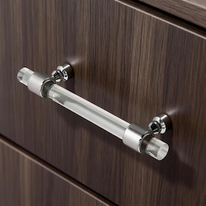 Puede incluir: Un tirador de armario de acrílico transparente y cromo. El tirador es cilíndrico con tapas de extremo cromadas y herrajes de montaje. El tirador está montado en un armario de madera marrón oscuro.