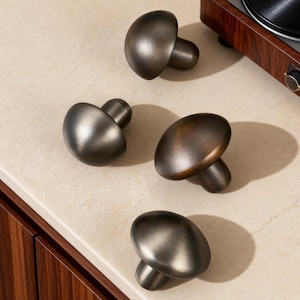 Könnte beinhalten: Vier bronzefarbene Schrankknöpfe in Pilzform. Die Knöpfe haben eine abgerundete Oberseite und einen zylindrischen Schaft. Die Oberfläche ist eine Mischung aus Dunkelbraun und Bronze mit einem leicht gealterten Aussehen. Die Knöpfe sind auf einer hellen Oberfläche angeordnet.