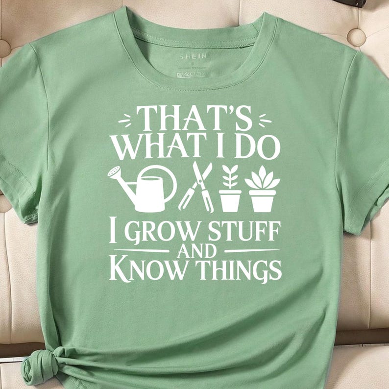 Puede incluir: Una camiseta de manga corta color verde salvia con el texto impreso en blanco THAT'S WHAT I DO I GROW STUFF AND KNOW THINGS, junto a iconos gr&aacute;ficos blancos de una regadera, tijeras de podar y dos plantas en maceta.