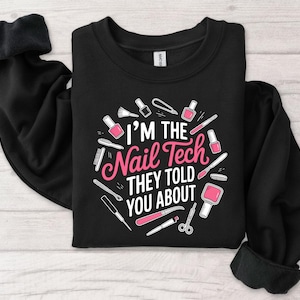 May include: Black crewneck sweatshirt with the text "I'M THE Nail Tech THEY TOLD YOU ABOUT" in pink and white. The design includes illustrations of nail polish bottles, brushes, and other manicure tools. The sweatshirt has long sleeves.