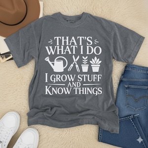 Pode incluir: Camiseta cinza com o texto branco "THAT'S WHAT I DO, I GROW STUFF AND KNOW THINGS". O design inclui um regador, tesouras e plantas em vasos. A camiseta é mostrada com jeans azul e tênis brancos.