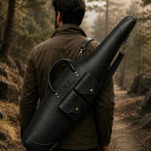 Könnte beinhalten: Ein schwarzes Gewehrfutteral aus Leder mit einem Tragegriff, einem verstellbaren Schultergurt und zwei Vordertaschen mit Messingdruckknöpfen. Das Futteral ist mit den Initialen M.F. geprägt und verfügt über Metallbeschläge für den Transport.