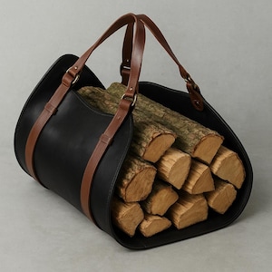 May include: A black leather log carrier with brown leather handles and straps. The carrier is filled with cut firewood, showcasing the natural wood grain and texture. The design is simple and functional.