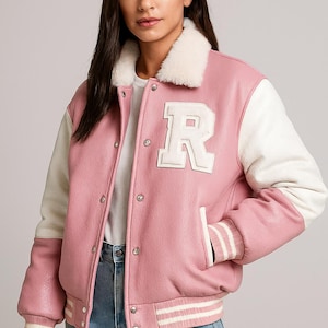 May include: A pink and white varsity jacket featuring a large white "R" on the chest. The jacket has a faux fur collar, white sleeves, and striped cuffs. The jacket is made of a smooth material and has a classic, sporty design.