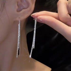 Puede incluir: Pendientes colgantes plateados con un aspecto brillante y reluciente. Los pendientes son largos y lineales, con un delicado diseño de cadena. Una mano sostiene los pendientes, mostrando su estilo elegante y llamativo.