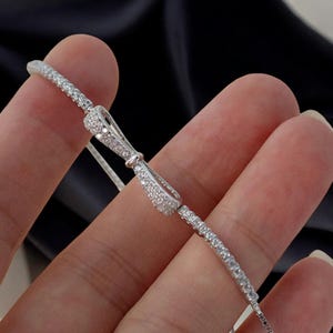 Puede incluir: Una delicada pulsera de plata con un elemento central en forma de lazo, incrustada con pequeñas piedras claras y brillantes. La cadena de la pulsera también está adornada con piedras similares, creando un brillo continuo. La joya se muestra sobre un fondo oscuro.