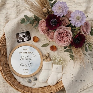 May include: A circular wooden-framed mirror with the text "Baby on the Way" and "Baby Smith April 202X" is displayed. The image features a sonogram, flowers, baby socks, and decorative items, creating a pregnancy announcement theme.