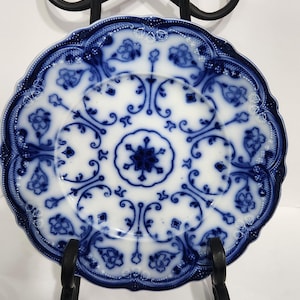 May include: A white and blue porcelain plate with a floral design, displayed on a black metal stand. The plate features a detailed blue pattern around the rim and center, showcasing a vintage aesthetic.