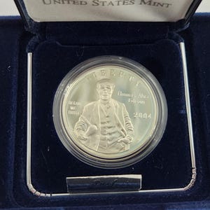 May include: A silver coin from the United States Mint, encased in a clear plastic holder and presented in a blue velvet box. The coin features a portrait of a man and the year 2004. The box is open, revealing the coin.