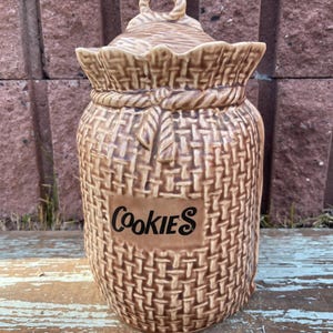 May include: A brown ceramic cookie jar with a woven basket design. The jar has a lid with a loop handle and the word "Cookies" in black lettering on the front. The jar is sitting on a wooden surface.