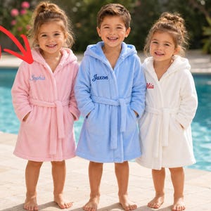 May include: Three plush hooded bathrobes in pink, light blue, and white, each featuring a tie waist, two front pockets, and a custom embroidered name on the chest. The pink robe displays the name Sophia, the blue robe Jaxon, and the white Mia.