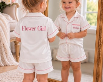 May include: A white short-sleeve pajama set with pink piping trim. The back of the shirt features the text Flower Girl in pink lettering. The front pocket of the shirt features the name Olivia in pink lettering. The shorts have an elastic waistband.