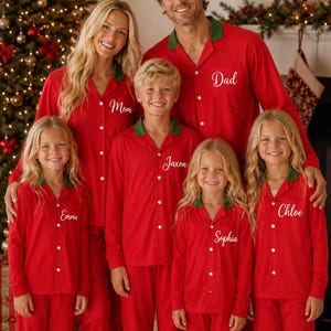May include: Matching red pyjama sets with green collars and white buttons. Each shirt features a white embroidered name on the chest pocket: Mom, Dad, Jaxon, Emma, Sophia, and Chloe. The long-sleeved tops and trousers are made of a solid red fabric.