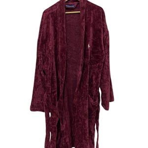Pode incluir: Robe de veludo bordô com gola xale, mangas compridas e dois bolsos frontais. O robe tem um cinto a condizer e um pequeno logótipo bordado no peito. Está pendurado num cabide de madeira.
