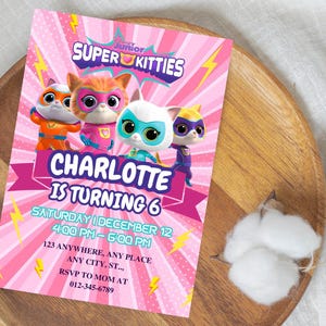 May include: A pink Super Kitties birthday party invitation featuring cartoon kittens in superhero costumes. The invitation includes the text "Charlotte is turning 6" and event details. The invitation is on a wooden tray.