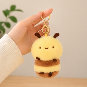 May include: A plush bee keychain featuring a soft, fuzzy yellow and brown striped body with small dark brown antennae, tiny arms, and legs. The character has a simple embroidered face with two small eyes and a curved mouth, attached to a gold metal ring.
