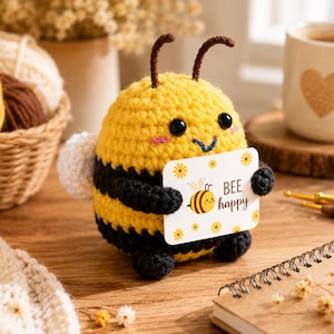 Può includere: Un peluche a forma di ape realizzato all'uncinetto, con strisce gialle e nere, occhi in plastica nera e due antenne. L'ape tiene in mano un cartoncino rettangolare bianco con una piccola illustrazione di un'ape, fiori gialli e il testo BEE happy.