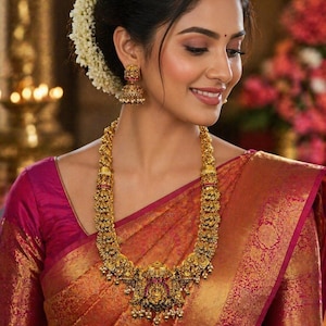 May include: A woman wearing a gold necklace and matching earrings. The necklace is long and ornate, with intricate detailing and red accents. The earrings are a similar design. She is wearing a pink and gold sari.