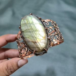 May include: A copper cuff bracelet featuring a large, oval-shaped labradorite gemstone as the centerpiece. The metal band is crafted with an intricate, openwork filigree design consisting of swirling patterns and small circular accents.