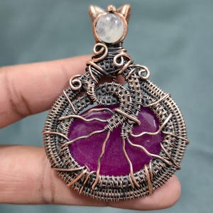 May include: A handcrafted pendant featuring a vibrant purple gemstone at its center, encased in intricate copper wire weaving. The pendant is topped with a small, round, white stone and has a decorative bail. The design showcases a blend of natural elements and artistic craftsmanship.