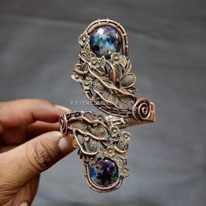 Puede incluir: Un brazalete de alambre de cobre enrollado con dos cabujones redondos de colores moteados y adornos florales de metal. El diseño incluye detalles de alambre en espiral y el texto RSJEWELLERYHOUSE impreso en el centro.