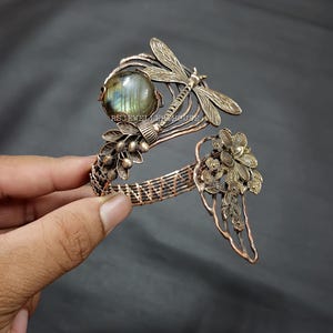 Könnte beinhalten: Ein kupferfarbenes, drahtgewickeltes Armband mit einem runden, irisierenden Labradorit-Edelstein, einem detaillierten Libellenmotiv und floralen Akzenten. Die Metallarbeit zeigt filigrane Blattmuster und den Text RSJEWELLERYHOUSE.