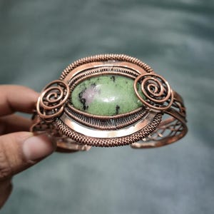 Op de afbeelding: Koperdraad gewikkelde armband met een ovale groene steen met rode insluitsels. De armband heeft ingewikkeld draadwerk en spiraalvormige accenten. Het metaal heeft een warme, roodbruine tint.