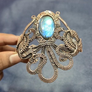 May include: A wire-wrapped cuff bracelet crafted from copper-toned metal wire, featuring an oval-shaped iridescent blue labradorite gemstone at the center. The intricate metalwork forms a series of swirling, tentacle-like loops and patterns.