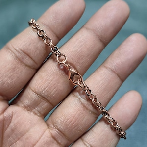 Op de afbeelding: Een koperkleurige ketting met een karabijnsluiting. De ketting is gemaakt van kleine, onderling verbonden ovale schakels. De ketting wordt gepresenteerd tegen een zachte, neutrale achtergrond.