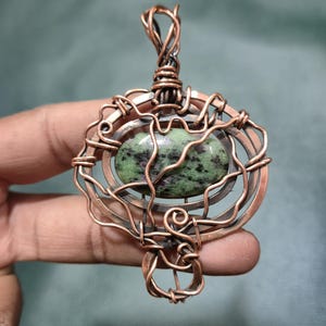 May include: A handcrafted copper pendant featuring a green and black oval stone. The stone is encased in a wire-wrapped design with intricate details. The pendant has a loop at the top for a chain. The overall design is rustic and artistic.
