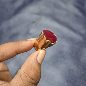 Op de afbeelding: Een gouden ring met een grote, ovaalvormige, dieprode edelsteen gezet in een dikke, gepolijste metalen band. De steen heeft een gefacetteerd oppervlak en wordt op zijn plaats gehouden door een klauwzetting in het goudkleurige metalen frame.