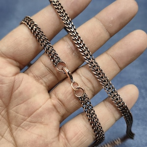 May include: A copper-toned chain necklace featuring a woven, textured link design. The chain is constructed with a repeating interlocking pattern and is secured with a small, circular lobster claw clasp mechanism made of the same metallic material.
