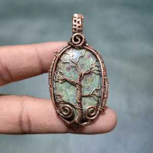 May include: Oval pendant featuring a green stone with a copper wire tree design. The tree has a detailed trunk and branches. The pendant is encased in a copper wire frame with decorative swirls. The pendant is held by a person's fingers.
