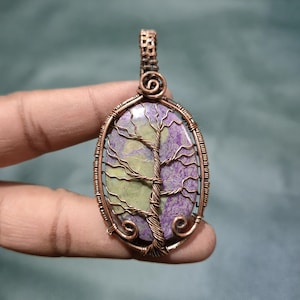 May include: Oval pendant with a copper wire frame and a tree design. The pendant features a stone with green and purple hues. The tree design is crafted with copper wire, adding a natural and artistic touch to the jewelry.