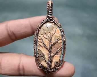 May include: Oval pendant featuring a tree design. The pendant has a copper-colored wire frame and a stone center with a tree-of-life design. The stone has tan and brown tones. The pendant is held by a person's fingers.