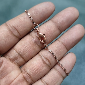 Puede incluir: Collar de oro rosa con una delicada cadena con un patrón de estrella repetido. El collar tiene un cierre de mosquetón. La cadena se sostiene en una mano sobre un fondo verde azulado.