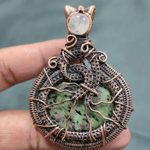 May include: A handcrafted pendant featuring a green and purple stone, intricately wrapped in copper wire. The pendant has a circular design with a tree-like pattern and a small, round, white stone at the top. The wirework is detailed and artistic.